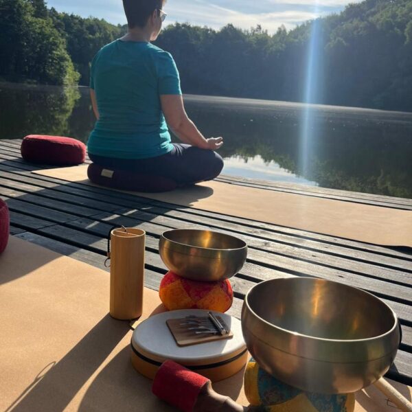 Klangschalen im Vordergrund, Daniela bereitet sich auf eine individuelle Yogastunde vor und blickt auf einen ruhigen See