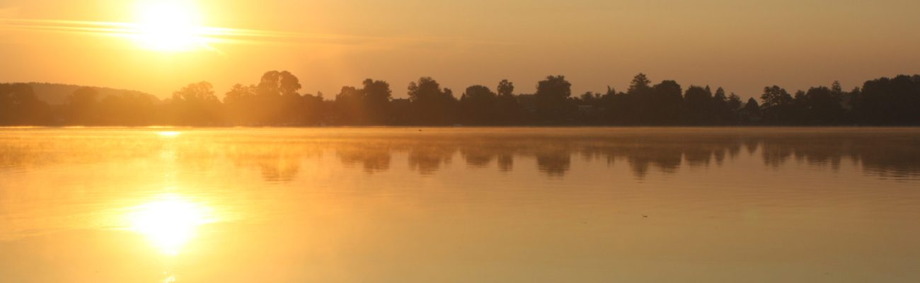 Einatmen. Ausatmen. Ankommen. Sonnenuntergang in warmen Gelb- und Orangetönen über einem ruhigen See mit Bäumen im Hintergrund – Naturidylle in Neuzelle