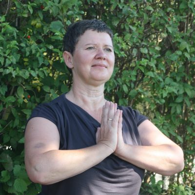 Daniela sitzt im Grünen und den Händen in Namaste-Haltung vor dem Herzen – Zeichen für Achtsamkeit, Dankbarkeit und innere Ruhe