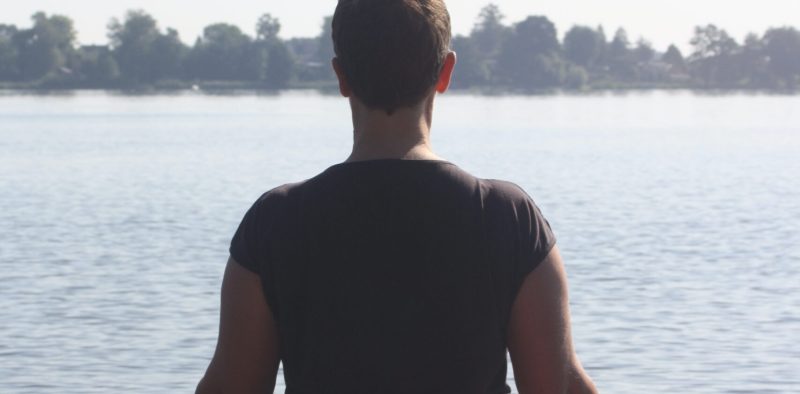Daniela sitzt im Schneidersitz auf einem Steg am Wasser, mit dem Rücken zum Betrachter – meditative Haltung in der Natur