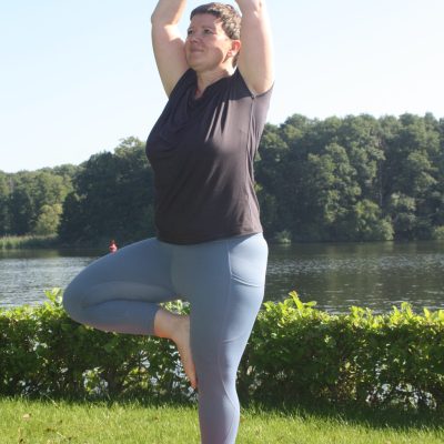 Daniela in der Baumstellung (Vrksasana) auf einer Wiese, im Hintergrund ruhiges Wasser – Balance und Naturverbundenheit
