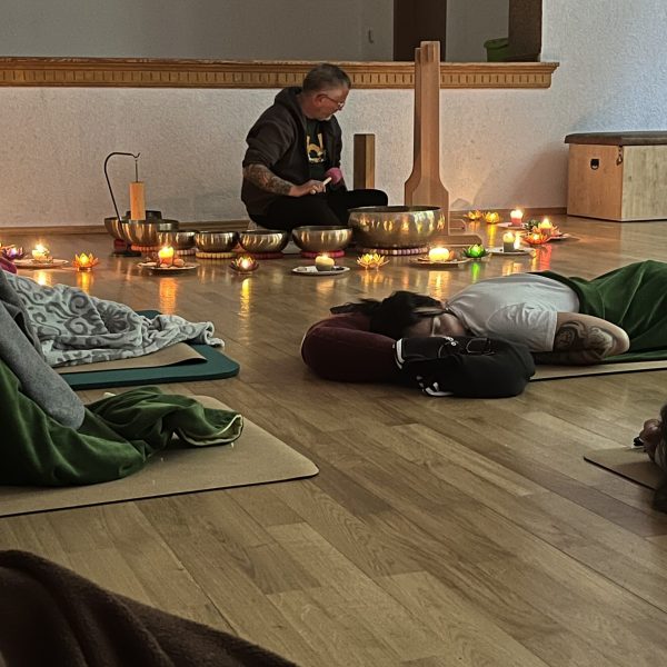 Ricardo umgeben von Klangschalen, im Hintergrund; im Vordergrund liegen Teilnehmer entspannt auf Yogamatten